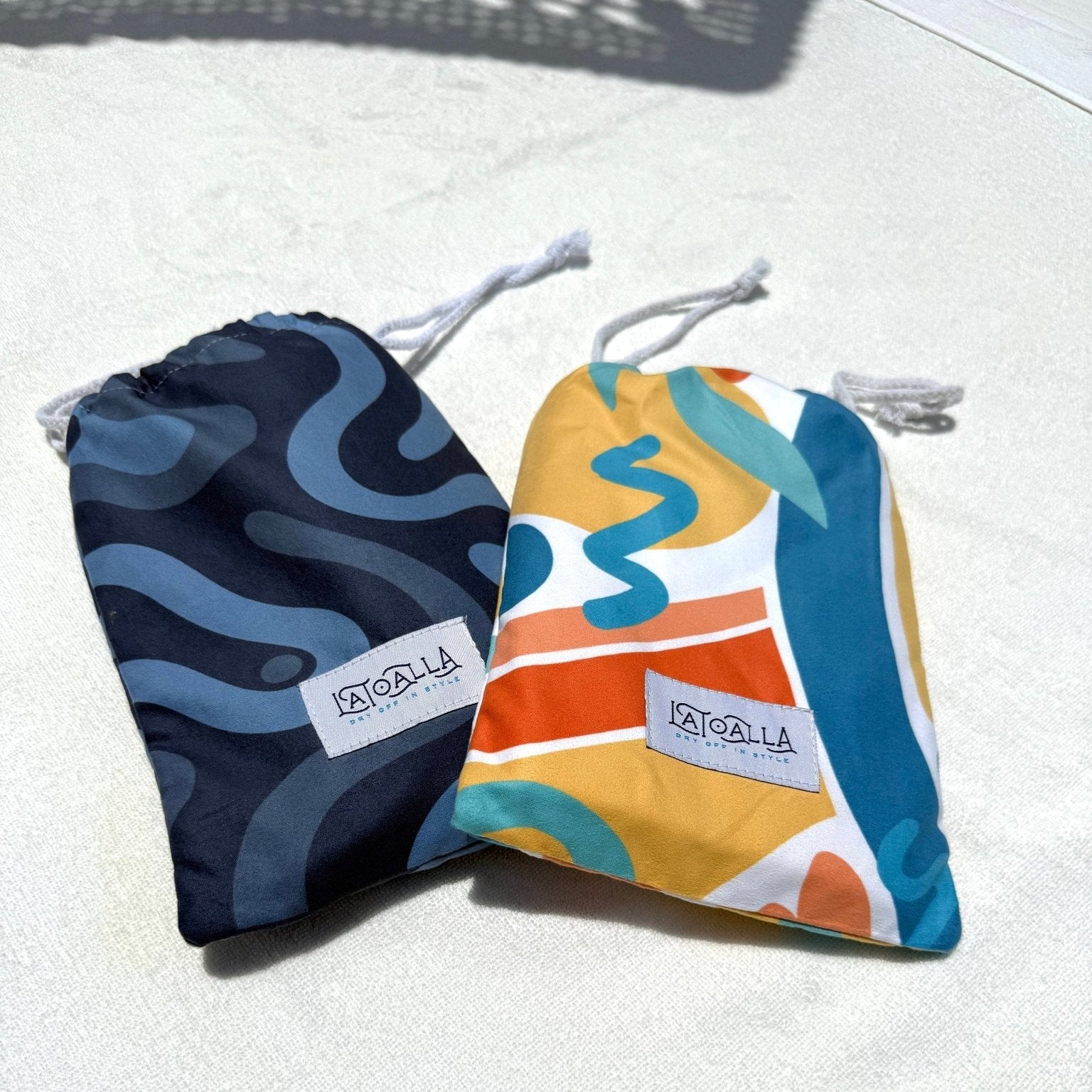 Two drawstring towel pouches from La Toalla, one navy blue with a light blue wavy pattern and the other yellow, orange, and blue with abstract shapes, displayed on a light textured surface.