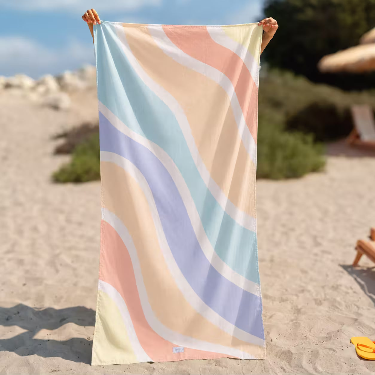 A rectangular beach towel with a pastel wavy stripe pattern in peach, light blue, lavender, and cream, held up by a person&