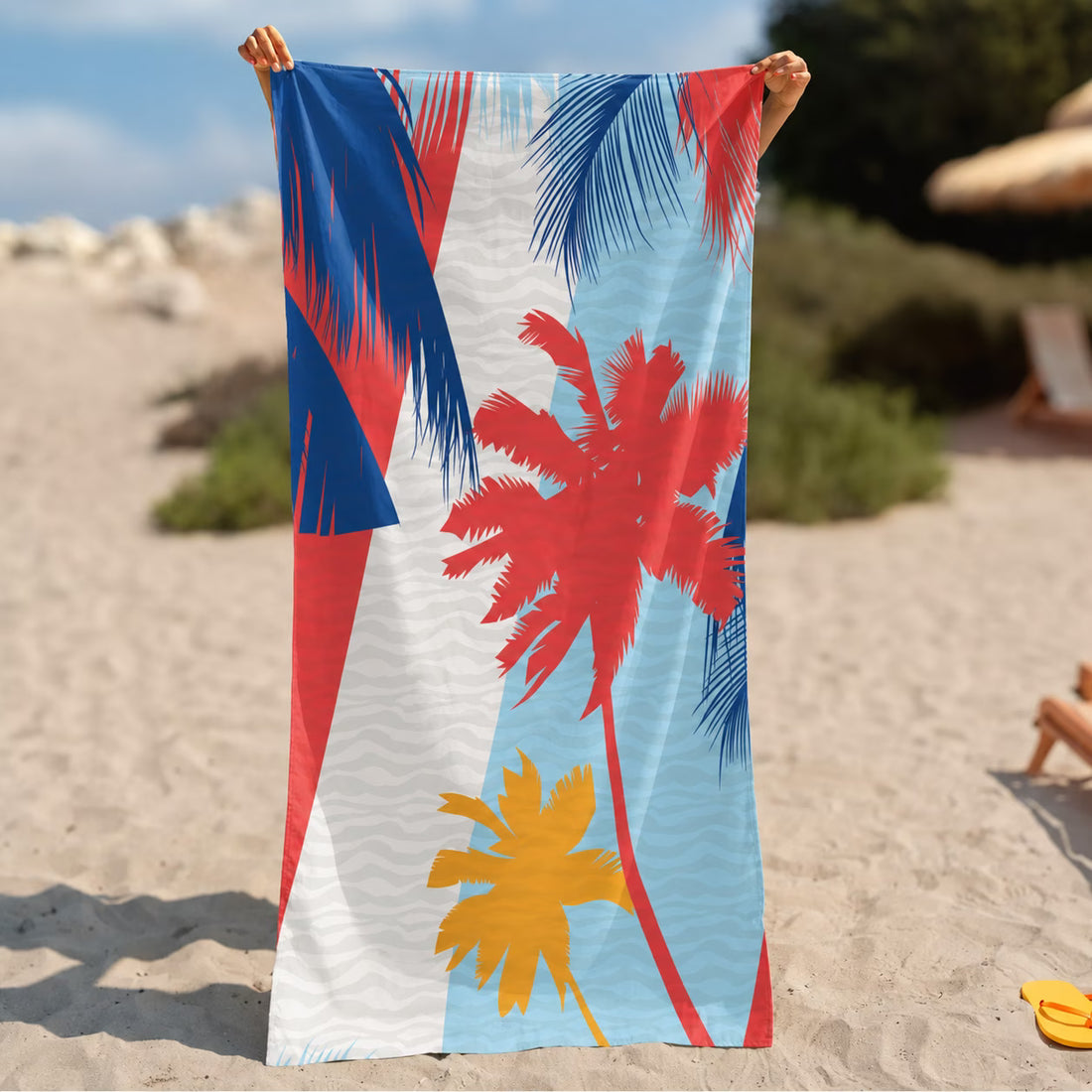 A beach towel with a tropical palm tree print in red, blue, yellow, and white, held up by a person on a sandy beach.