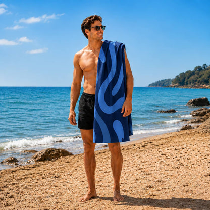 A shirtless man wearing sunglasses stands on a sandy beach with the ocean and blue sky behind him. He is holding a blue patterned towel draped over his shoulder.