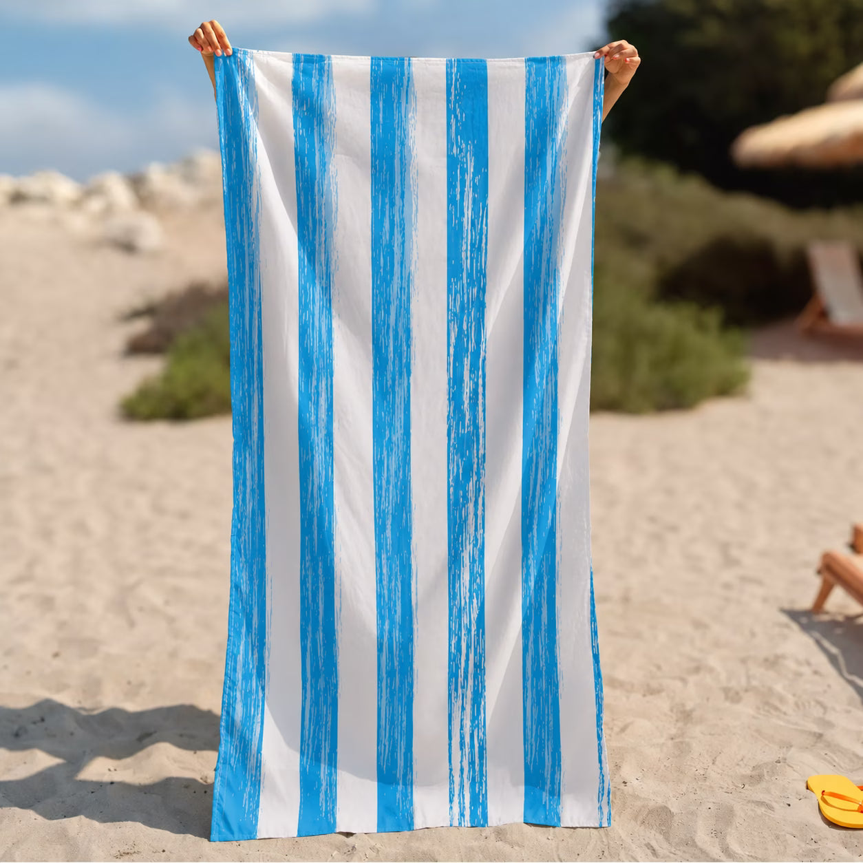A person holds up a large beach towel from La Toalla with vertical blue and white stripes on a sandy beach.
