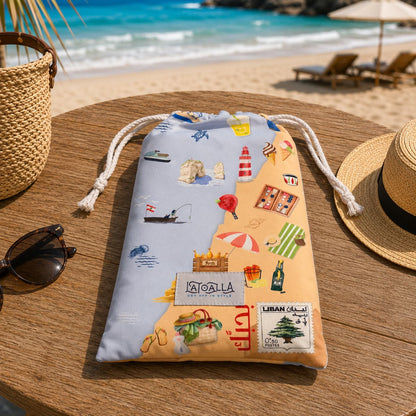 A drawstring beach towel bag from La Toalla with a colorful travel-themed print, displayed on a wooden table at a beach with lounge chairs, sunglasses, and a straw hat nearby.