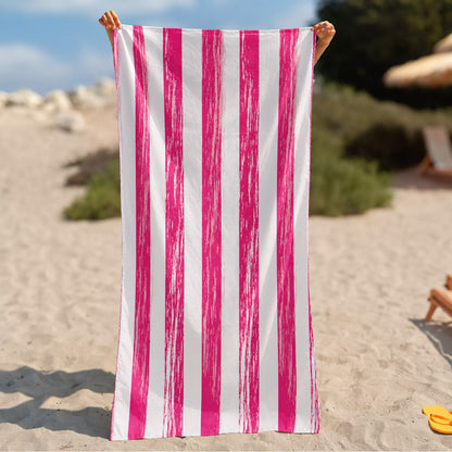 A beach towel with bold vertical pink and white stripes is held up by two hands on a sandy beach.