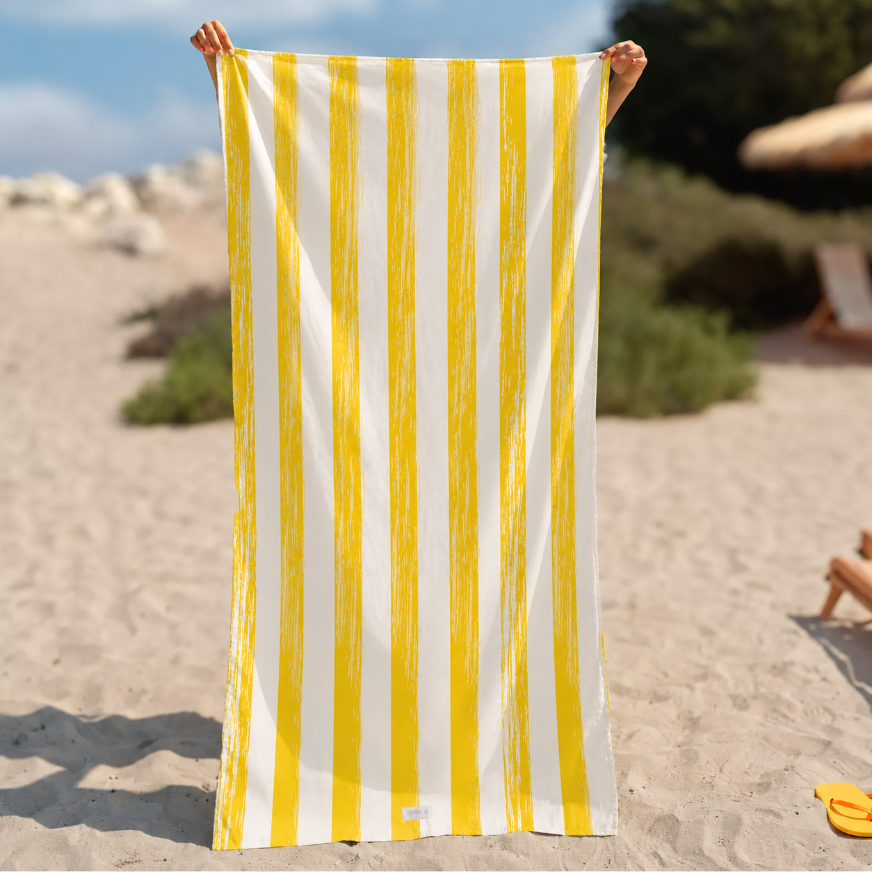A person on a sandy beach holds up a large beach towel from La Toalla l with vertical yellow and white stripes.