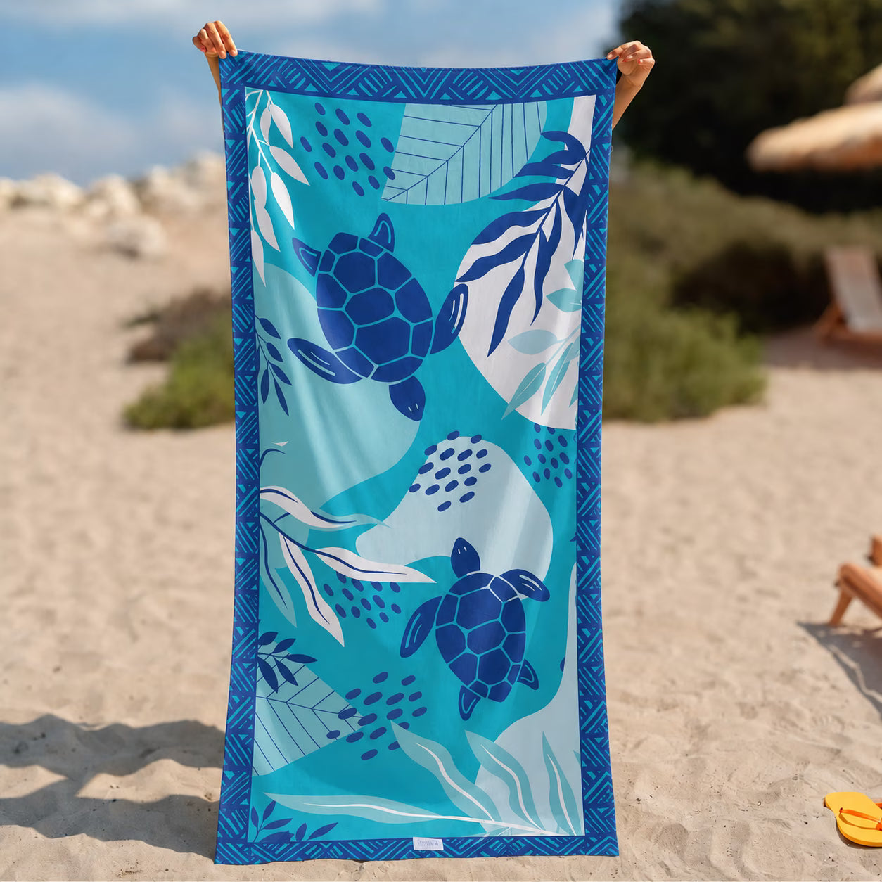 A person holds up a rectangular beach towel on a sandy beach. The towel features a blue and teal ocean-themed pattern with sea turtles, leaves, and waves, and has a decorative blue border from La Toalla