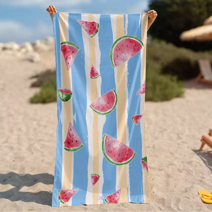 A person holds up a large beach towel on a sandy beach. The towel features a light blue background with vertical cream stripes and watermelon slice illustrations in pink, red, and green. Microfiber Beach Towel from La Toalla